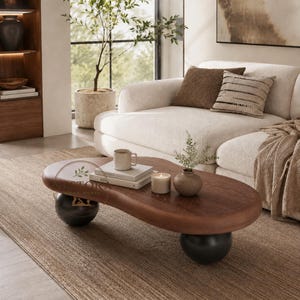 May include: A kidney-shaped coffee table with a dark brown wood grain top and two large, smooth black spherical legs. The table features a subtle logo with a leaf icon and the text NESTOKA WOOD WORKS printed on the surface.