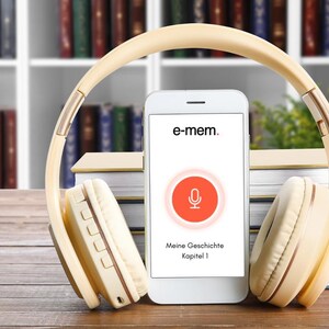 以下が含まれることがあります： e-memというテキスト、マイクアイコン、Meine Geschichte Kapitel 1という文字が表示された白いスマートフォン。その上にベージュのオーバーイヤーヘッドホンが置かれています。画面には円形のオレンジ色の録音インターフェースが表示されています。