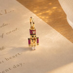 May include: A gold-toned metal charm shaped like a nutcracker, featuring magenta glitter enamel details on the hat and sleeves, a small clear stone star on the hat, and two small white spherical beads representing the feet, attached to a gold ring.