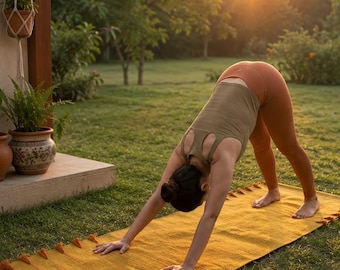May include: A rectangular mustard yellow yoga mat featuring decorative tassels along the edges. The mat is textured and laid out on a grassy surface, providing a base for a person performing a downward-facing dog yoga pose in athletic wear.