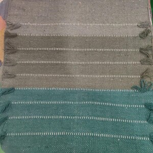 May include: A rectangular woven textile runner featuring a color-blocked design with a white top section, followed by horizontal bands of sage green and dark forest green. The edges are accented with small, matching green fringe tassels.