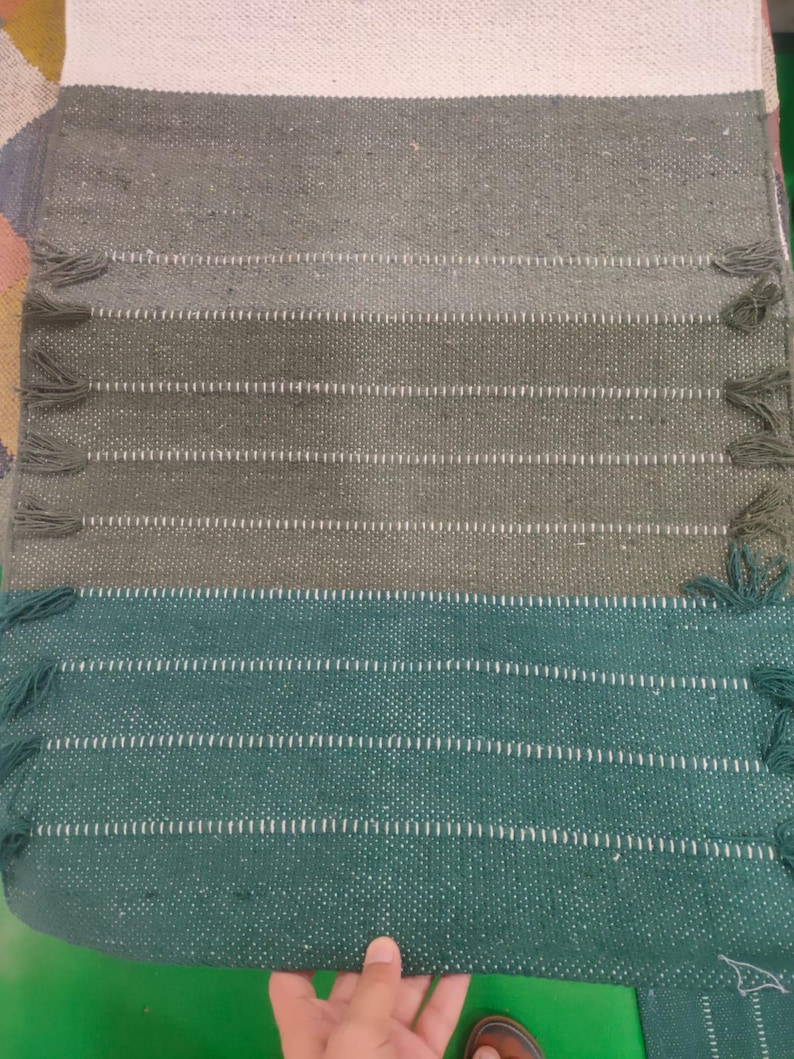 May include: A rectangular woven textile runner featuring a color-blocked design with a white top section, followed by horizontal bands of sage green and dark forest green. The edges are accented with small, matching green fringe tassels.