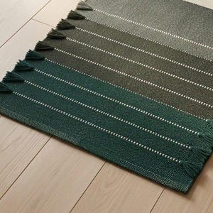 May include: A rectangular woven rug featuring a gradient of green shades, transitioning from dark forest green to lighter olive tones. The rug is accented with thin white horizontal stripes and finished with small tassels along the edges.