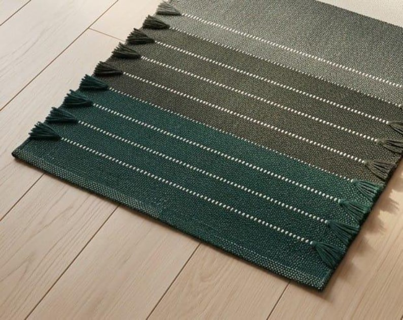 May include: A rectangular woven rug featuring a gradient of green shades, transitioning from dark forest green to lighter olive tones. The rug is accented with thin white horizontal stripes and finished with small tassels along the edges.