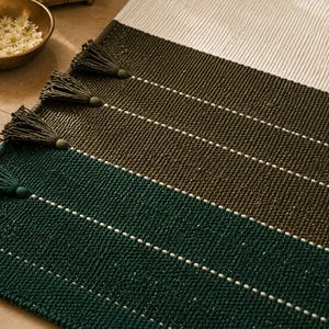 May include: A rectangular woven rug featuring horizontal color-blocked sections in cream, olive green, and dark teal. The rug is accented with thin white horizontal stripes and matching tassels attached along the side edges of the fabric.