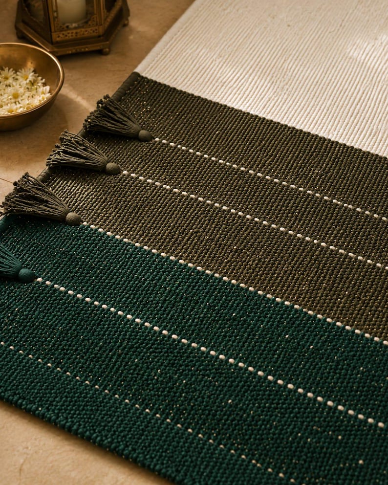 May include: A rectangular woven rug featuring horizontal color-blocked sections in cream, olive green, and dark teal. The rug is accented with thin white horizontal stripes and matching tassels attached along the side edges of the fabric.