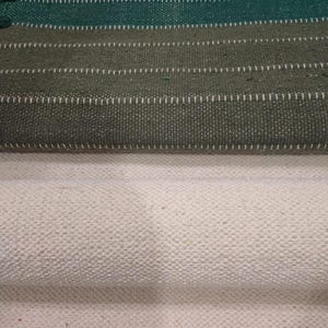 May include: A woven textile fabric featuring a top section with horizontal stripes in dark green and olive green, accented by thin white stitched lines. The bottom section consists of a solid, textured off-white fabric with a visible weave.