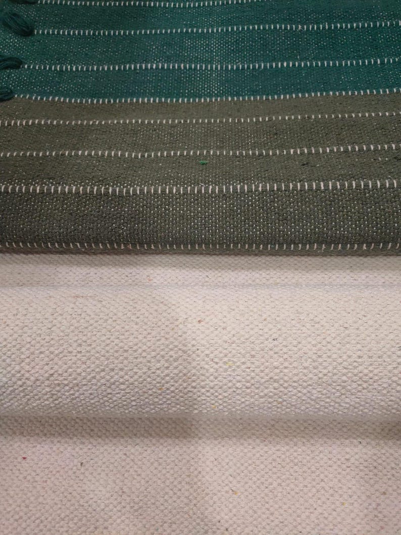 May include: A woven textile fabric featuring a top section with horizontal stripes in dark green and olive green, accented by thin white stitched lines. The bottom section consists of a solid, textured off-white fabric with a visible weave.