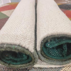 May include: Two cylindrical rolls of textile material placed side by side. The outer surface of the rolls is a textured off-white fabric, while the inner core reveals a deep forest green color with a visible woven structure and finished edges.