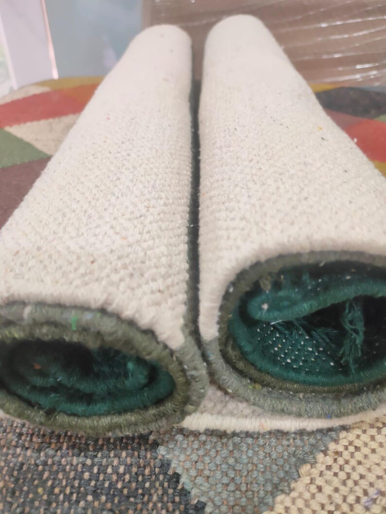 May include: Two cylindrical rolls of textile material placed side by side. The outer surface of the rolls is a textured off-white fabric, while the inner core reveals a deep forest green color with a visible woven structure and finished edges.