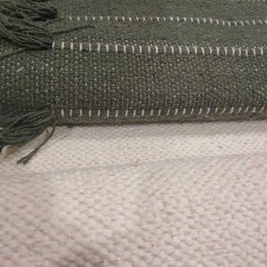 May include: A woven textile featuring horizontal stripes in shades of dark green and olive green, accented with thin white stitching lines and short fringe details along the edges, layered over a solid off-white textured fabric base.