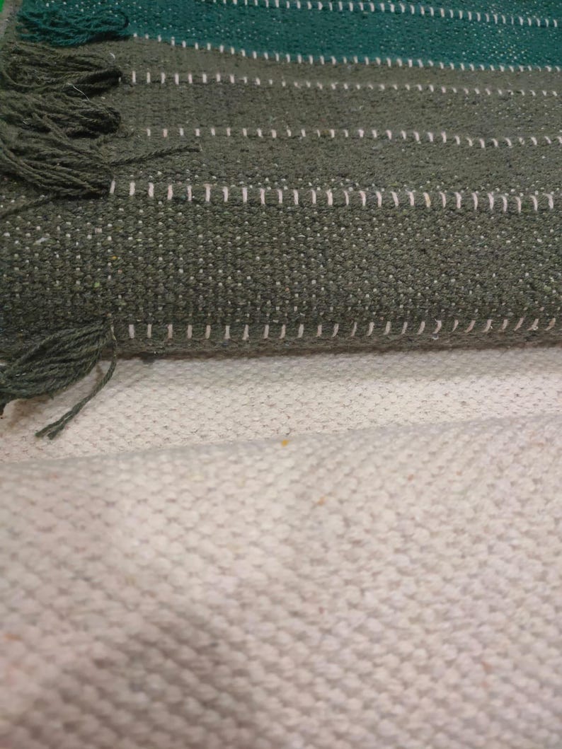 May include: A woven textile featuring horizontal stripes in shades of dark green and olive green, accented with thin white stitching lines and short fringe details along the edges, layered over a solid off-white textured fabric base.