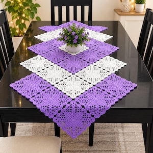 Peut inclure: Un chemin de table en crochet rectangulaire composé de motifs carrés alternés violets et blancs disposés en losange. Le chemin de table présente un design en dentelle ajourée complexe et est centré avec un petit napperon carré en crochet blanc.