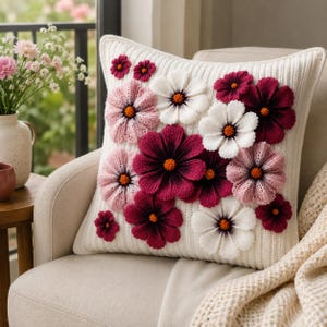 May include: A square white knit throw pillow featuring a textured ribbed pattern and multiple three-dimensional crocheted flowers in shades of pink, dark magenta, and white, each with a small orange center, arranged across the front surface.