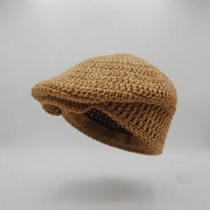 May include: A tan crochet flat cap featuring a textured, woven yarn construction with a rounded crown and a short, stiffened brim. The hat displays a consistent, dense stitch pattern throughout, creating a structured yet flexible form.
