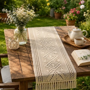 Op de afbeelding: Een crèmekleurige gehaakte tafelloper met een geometrisch ruitpatroon en franjes. De loper ligt op een houten tafel naast een glazen vaas met witte bloemen, een kleine kaarshouder, een theepot, een kopje en een boek.