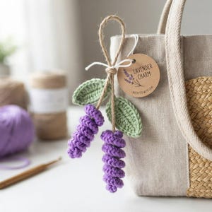 May include: A beige linen and woven straw tote bag featuring a hanging crochet lavender ornament. The ornament consists of two purple spiral-stitched lavender sprigs with green leaves, tied with twine and a round tag labeled Lavender Charm.