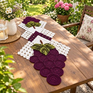 Pode incluir: Um caminho de mesa de crochê com dois cachos de motivos circulares roxo-escuros que lembram uvas, acentuados com folhas verdes texturizadas e unidos por painéis de renda branca vazada com um padrão geométrico.