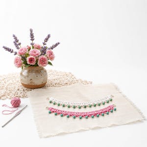 May include: Two curved crochet lace collars, one white and one pink, each featuring a row of small green beads along the bottom edge. A small ball of pink yarn and a metal crochet hook are positioned to the left of the lace pieces.