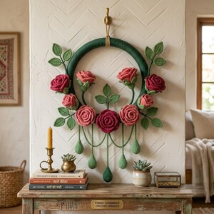 May include: A circular wall hanging featuring a dark green yarn-wrapped hoop adorned with crocheted roses in shades of red and pink, accompanied by green crocheted leaves and dangling teardrop-shaped accents hanging from the bottom of the wreath.