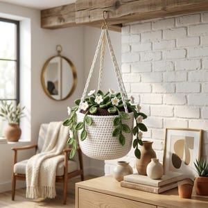 Puede incluir: Una maceta colgante de ganchillo blanco que contiene una planta con hojas de color verde oscuro y pequeñas flores blancas. La maceta está suspendida por un cordón de ganchillo blanco a juego de una viga de madera. La planta tiene enredaderas.