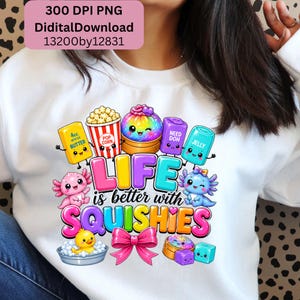 May include: A white sweatshirt featuring a colorful graphic print with the text "LIFE is better with SQUISHIES". The design includes cartoon characters of popcorn, butter, jelly, and axolotls, along with a yellow duck in a tub and a pink bow.