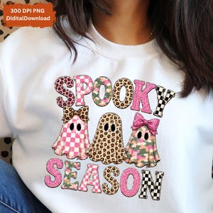 Könnte beinhalten: Ein weißes Sweatshirt mit einer Grafik von drei Geistern in verschiedenen Mustern, darunter Leopardenmuster, rosa Karomuster und Camouflage. Der Text "SPOOKY SEASON" ist in einer Mischung aus rosa Glitzer, Leopardenmuster und Karos gedruckt.