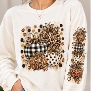 May include: A cream-colored long-sleeve sweatshirt featuring a central graphic of four pumpkins with leopard, buffalo plaid, and polka dot patterns. The left sleeve displays a vertical column of four smaller pumpkins with matching patterns.