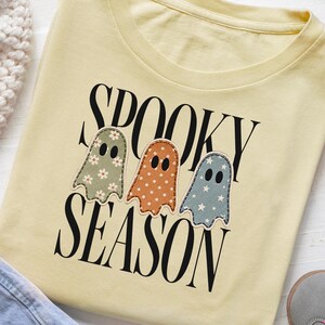 Könnte beinhalten: Ein blassgelbes T-Shirt mit dem schwarzen Schriftzug SPOOKY SEASON. Das Design zeigt drei geisterförmige Aufnäher: einen salbeigrünen mit weißen Gänseblümchen, einen orangefarbenen mit weißen Punkten und einen blauen mit weißen Sternen.