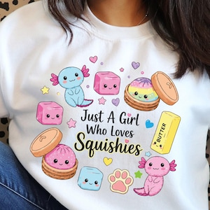 Może przedstawiać: Biała bluza z grafiką z napisem "Just A Girl Who Loves Squishies" otoczona ilustracjami dwóch aksolotli, dwóch bułeczek na parze, kostki masła, trzech kolorowych kostek, odcisku łapy oraz serduszek.