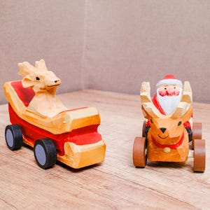May include: Two hand-carved wooden figurines on wheels. One is a tan reindeer pulling a red and tan sleigh. The other is a tan reindeer with a Santa Claus figure riding on its back, featuring a red hat, white beard, and red suit with black wheels.
