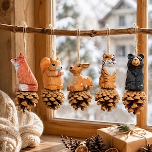 May include: Five hand-carved wooden animal ornaments hanging from a branch, each attached to a natural pinecone. The figures include a red fox, a light brown squirrel, a tan deer, a striped tiger, and a black bear, all suspended by twine.