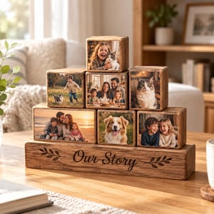 Op de afbeelding: Een houten displaystandaard met de gegraveerde tekst "Our Story" en twee bladiconen, die een piramide van zes houten fotoblokken ondersteunt. Elk blok bevat een afgedrukte foto van mensen en huisdieren, waaronder een kind met een kat en een hond.