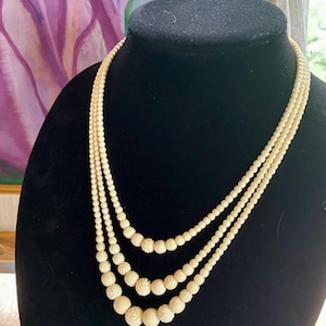 May include: A three-strand necklace featuring off-white, cream-colored beads of varying sizes. The strands are composed of smooth, round beads interspersed with smaller, spherical beads that have intricate, carved floral rose patterns.