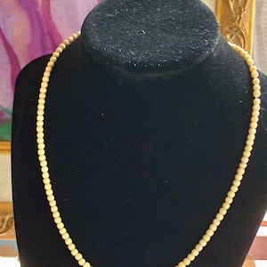 May include: A single strand necklace composed of small, uniform, cream-colored spherical beads. The beads are strung together in a continuous circular pattern, creating a simple and consistent design that rests evenly around the neck form.