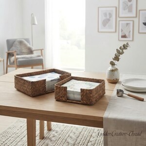 May include: Two rectangular woven seagrass baskets holding stacks of paper napkins. The baskets feature a natural brown texture and open-front design. They are placed on a light wood table alongside a ceramic vase with a branch and white plates.