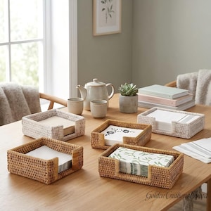 May include: Four rectangular woven rattan napkin holders in natural and whitewashed finishes, each containing a stack of paper napkins. The holders are arranged on a wooden table alongside a ceramic teapot, two cups, a small potted succulent, and books.