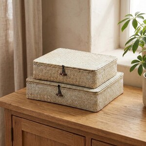 May include: Two rectangular woven seagrass storage boxes stacked on top of each other. Each box features a hinged lid secured with a black elastic cord and a small dark brown wooden toggle button centered on the front edge of the lid.