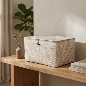May include: A rectangular storage box with a woven texture in light beige and white tones. The box features a matching lid secured by a small dark brown wooden toggle button and a thin cord loop closure, creating a simple and functional design.