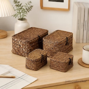 May include: A set of four rectangular woven storage baskets made from dark brown natural fibers. Each basket features a hinged lid secured with a black elastic cord and a small wooden toggle button closure. The baskets vary in size and depth.
