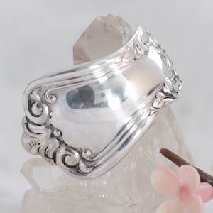 May include: Silver ring made from a vintage silverware spoon with an ornate design. The ring is curved and has a polished finish.