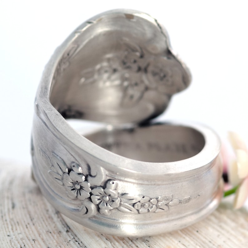 May include: A silver ring made from a repurposed spoon with floral designs. The ring is open and has a wide band.