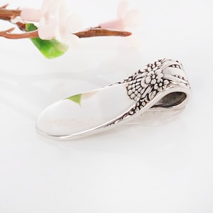 May include: A silver spoon pendant with a floral design. The spoon is curved and has a smooth, polished surface. The floral design is intricate and detailed.