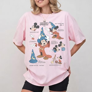 Pode incluir: Uma t-shirt rosa claro com uma impressão gráfica do Mickey Mouse como o aprendiz de feiticeiro. O design inclui várias ilustrações da personagem, texto instrutivo e diagramas que detalham técnicas de animação do filme de 1940.