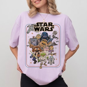 May include: A light purple short-sleeve t-shirt featuring a graphic print of Muppet characters dressed as Star Wars figures. The design includes the text STAR WARS at the top, with Kermit the Frog, Miss Piggy, Fozzie Bear, and other characters depicted in a retro-style frame.