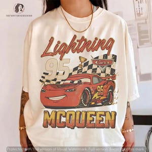 May include: An off-white short-sleeve t-shirt featuring a graphic of a red race car with the number 95. The design includes a checkered flag, a trophy, and the text Lightning 95 MCQUEEN. The print has a distressed, vintage-inspired aesthetic.