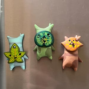 May include: Three small, sculpted figures with smooth surfaces. The left figure is light blue with a green leaf-shaped face. The centre figure is light green with a dark green circular face. The right figure is tan with an orange leaf-shaped face.