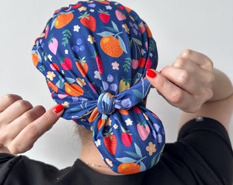 Op de afbeelding: Een blauwe stoffen hoofdbedekking met een patroon van aardbeien, sinaasappels, bosbessen en bloemen. De stof is aan de achterkant van het hoofd in een knoop geknoopt.