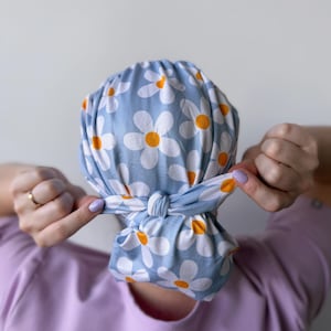 Op de afbeelding: Een blauw-witte scrubcap met madeliefjesprint is in een knoop gelegd. De cap heeft een lichtblauwe achtergrond met witte madeliefjes en oranje centra. De cap wordt vastgeknoopt door een persoon die een lichtpaars shirt draagt.