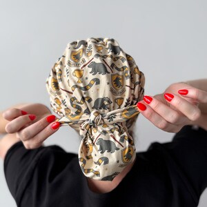 Op de afbeelding: Een beige scrub cap met een herhalend patroon van gele en zwarte sjaals, dassen en de tekst "Hufflepuff". De cap is achteraan geknoopt. De persoon draagt een zwarte top.